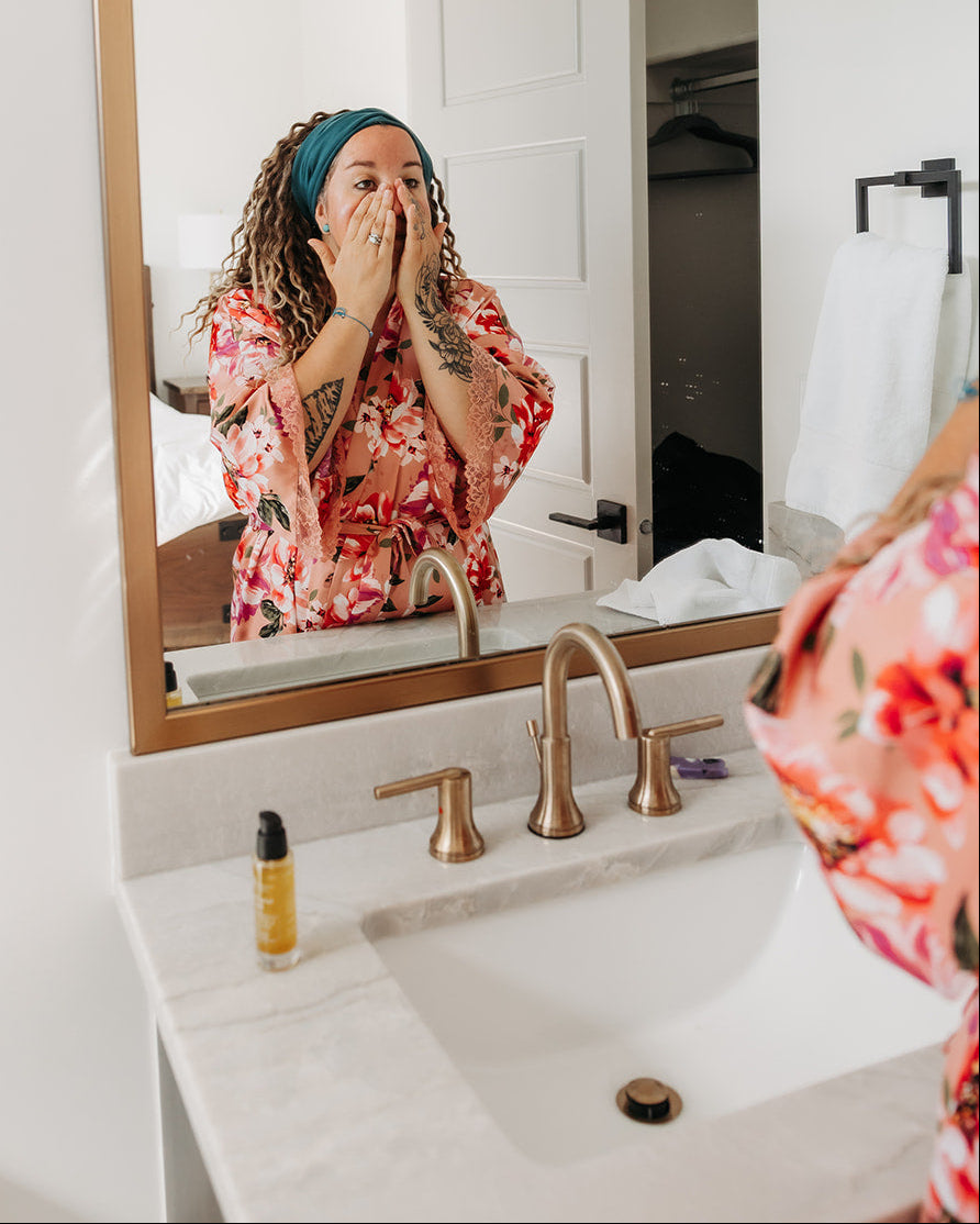 A woman in a red and pink floral robe stands in front of a bathroom mirror, touching her face. The mirror has a gold frame and reflects her hair and the bathroom interior.