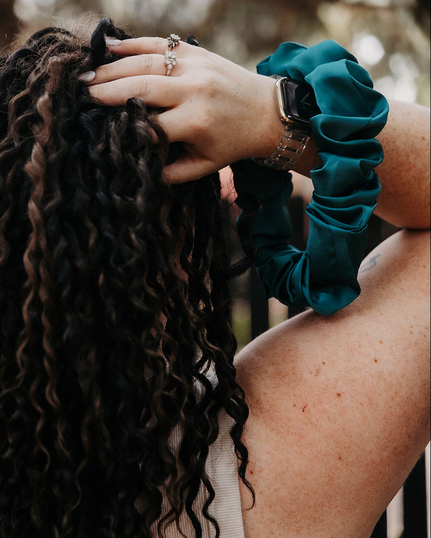 A close-up lifestyle shot of a person with long, dark, tightly curled hair from behind, wearing a dark teal scrunchie on one wrist, set against a blurred outdoor background.