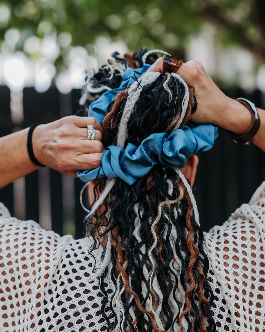 A person outdoors with long, curly black, brown, and white hair gathered into a messy bun, wearing a blue satin scrunchies. The person is seen from behind, wearing a white open-knit top, with a blurred fence and green foliage in the background.