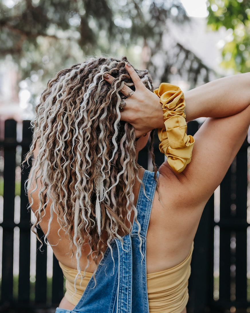 A woman with long, curly blonde hair extensions seen from behind, wearing denim overalls and a beige crop top, holding her hair up with a yellow scrunchie in front of a black fence and trees.