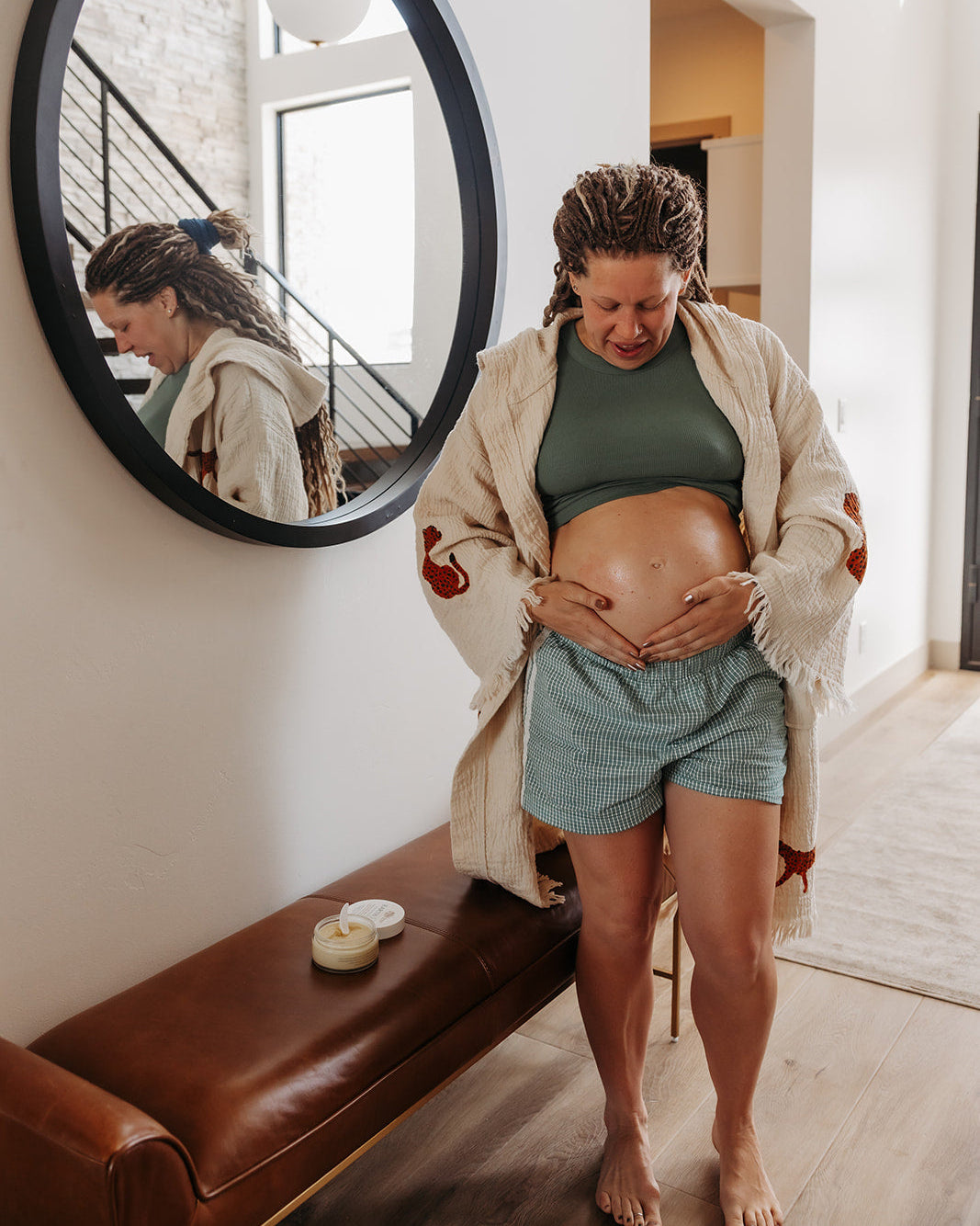 A pregnant woman stands barefoot in a bright room, wearing a cream fringed cardigan, a dark green crop top, and light blue shorts, applying Earth Butter on her baby bump. A large black-framed oval mirror on the wall reflects her back, and a brown leather bench sits below.