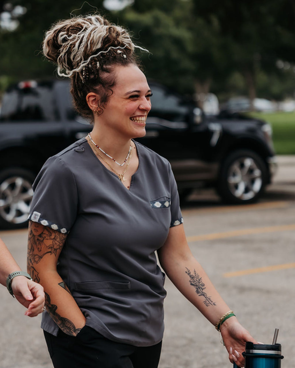 Person wearing Venus Long Braid-in Hair Extensions and a gray shirt with tattoos, holding a drink, in a parking lot.