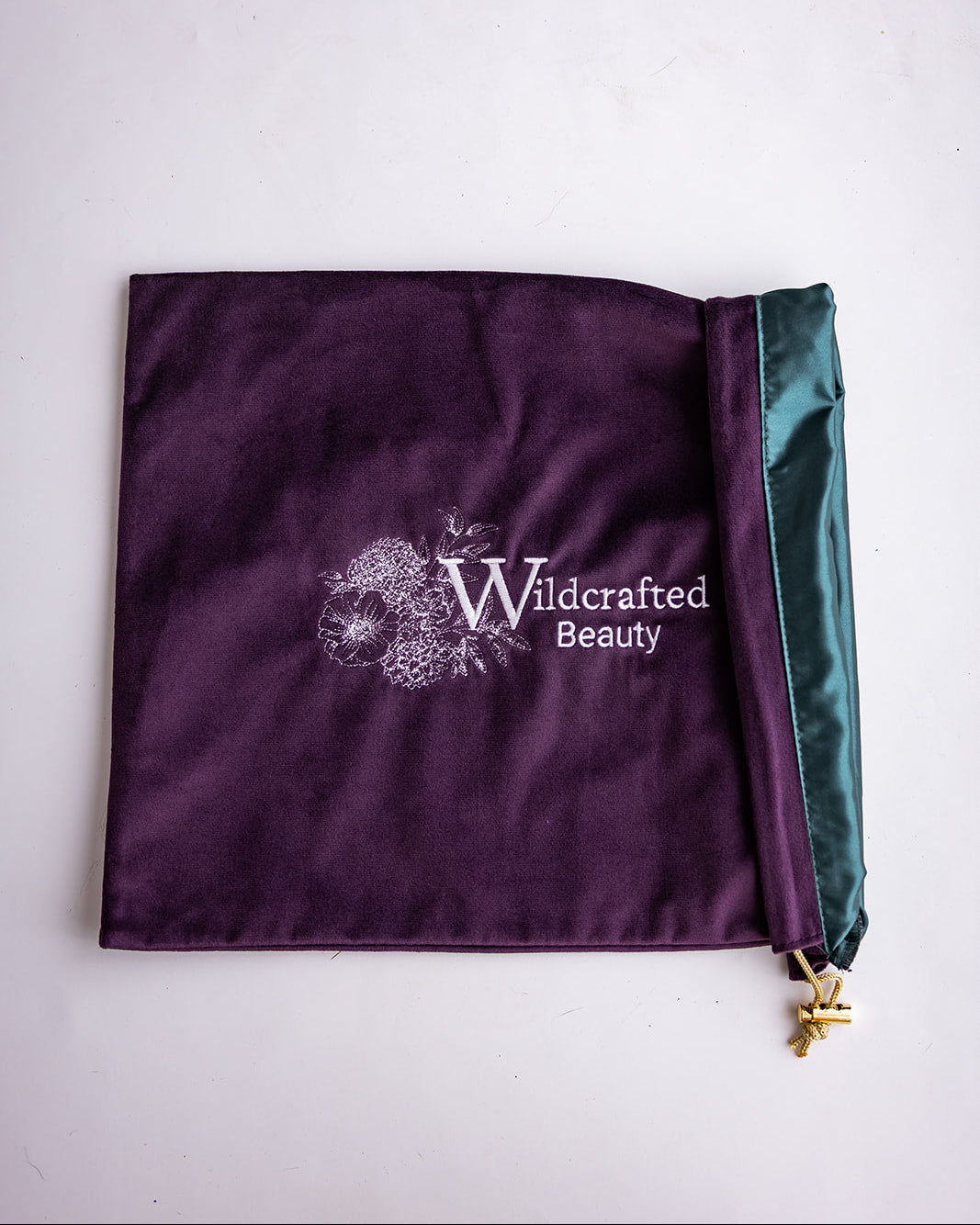 A dark purple velvet cosmetic pouch with a green satin zipper trim and a gold pull tab, featuring the white embroidered logo 'Wildcrafted Beauty' on the front, laid flat on a white surface.