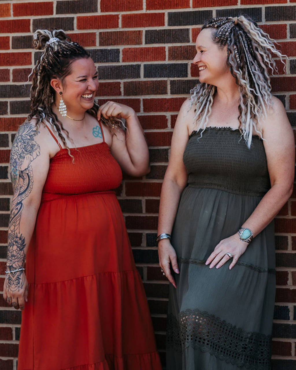 Two women wearing SHORTIES™ Braid-in Extensions standing against a brick wall, one in a red dress and the other in a dark green dress.