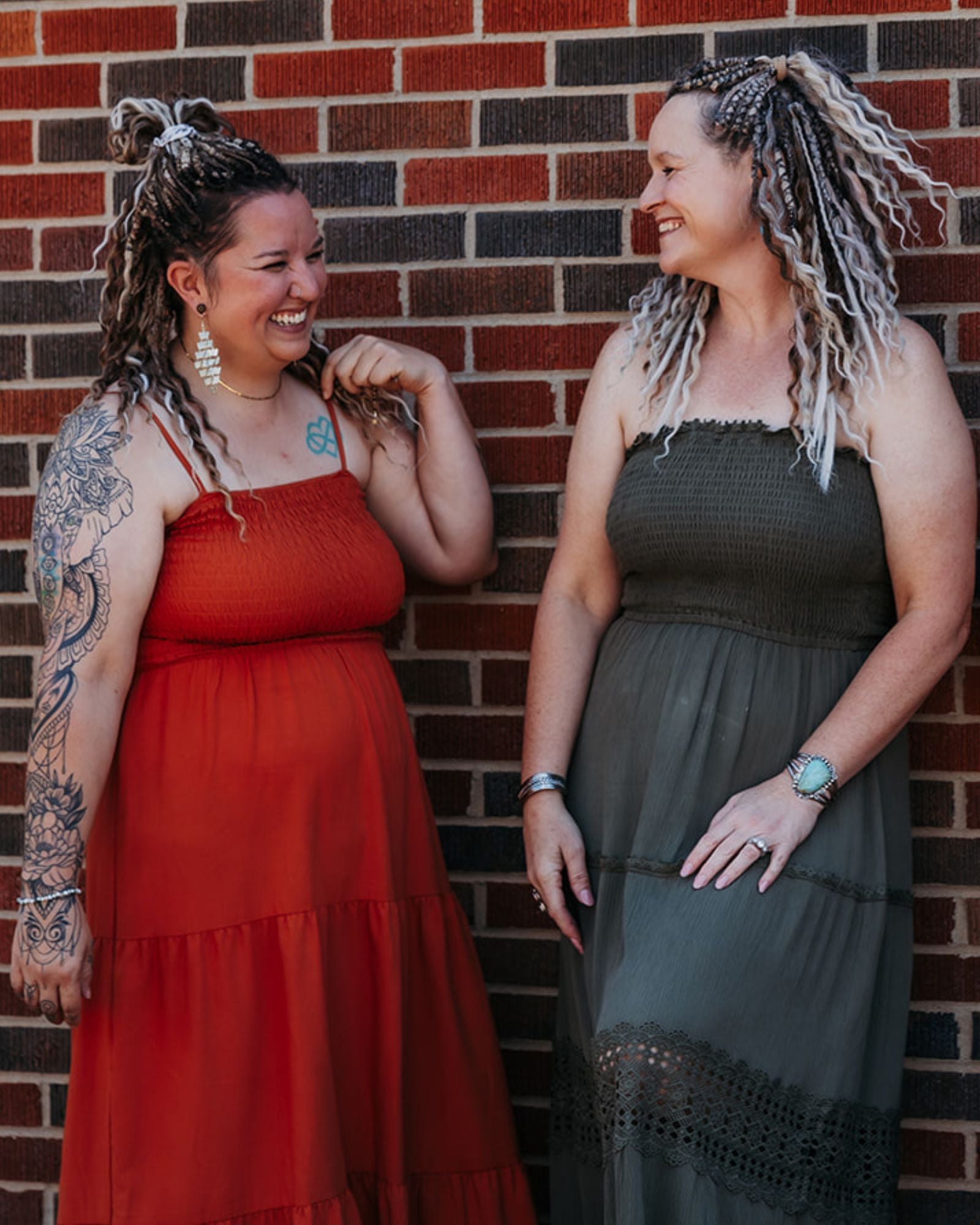 Two women wearing SHORTIES™ Braid-in Extensions standing against a brick wall, one in a red dress and the other in a dark green dress.