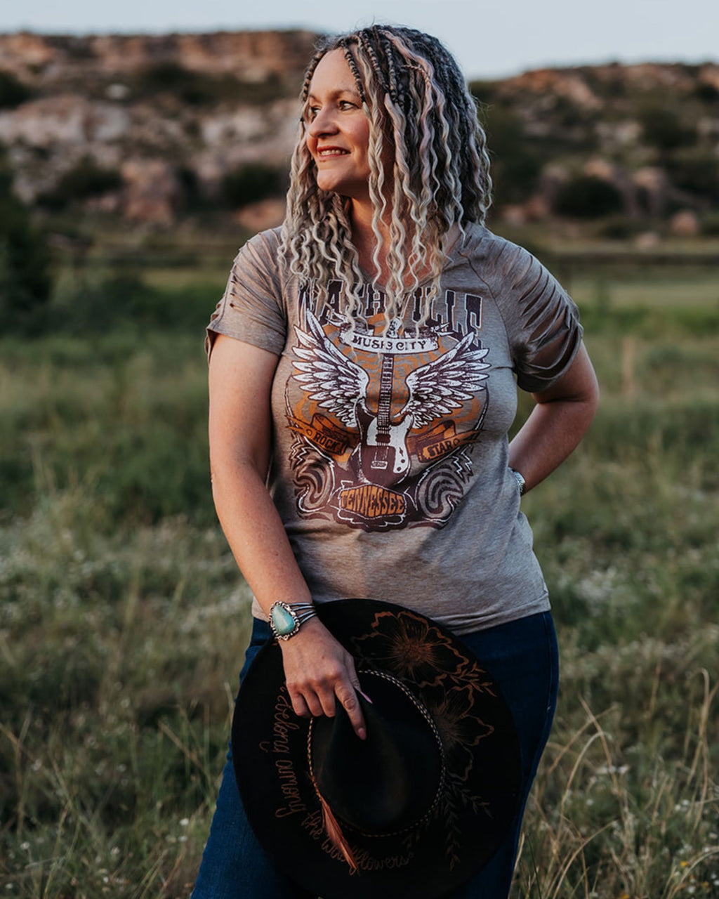 Woman wearing SHORTIES™ Braid-in Extensions and a graphic t-shirt holding a hat, standing in a field.