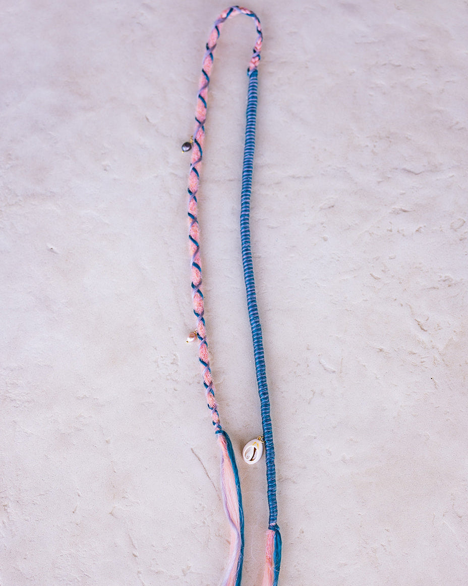 A handmade bohemian-style straight pink dread with blue braided wrap fiber, a small shell charm, and pearl charms, laid flat on a textured off-white surface.