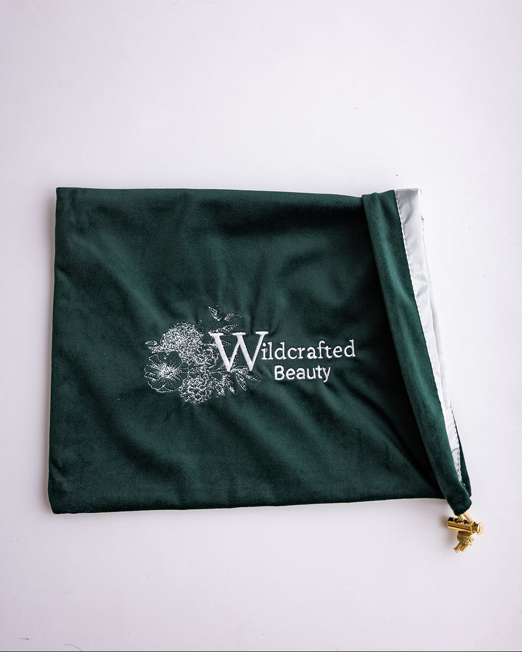 A dark forest green velvet drawstring pouch with a white 'Wildcrafted Beauty' logo printed on the front, shown on a light off-white background.