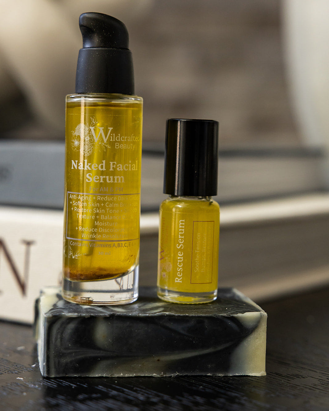 Two skincare bottles from Wildcrafted Beauty, including a larger Naked Facial Serum bottle and a smaller Rescue Serum bottle, displayed on a black and white marbled soap block.