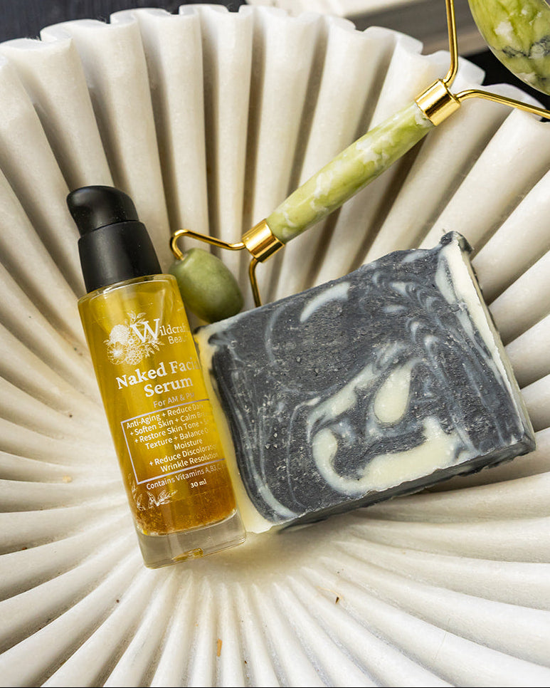 A small bottle of golden facial oil and a marbled gray-and-white soap bar are arranged on a white scalloped dish with a green-and-gold facial roller resting behind them on a dark surface.