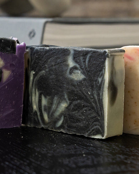 Black and White swirled soap next to a purple bar and beige bar on a dark wood table with a blurred background