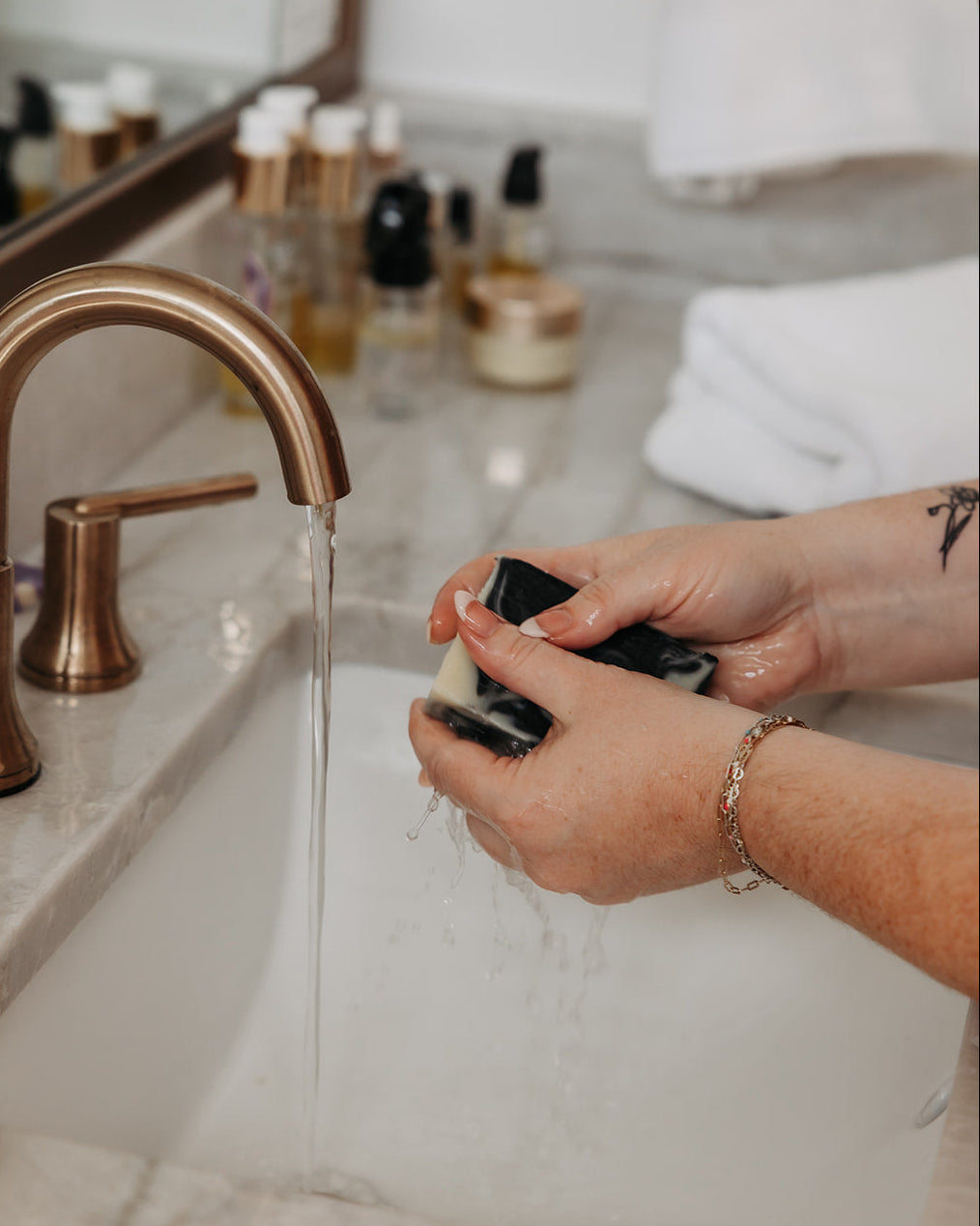 A close-up lifestyle shot of a person's hands holding a black and white marbled facial cleansing bar under running water from a matte bronze faucet in a marble bathroom sink.