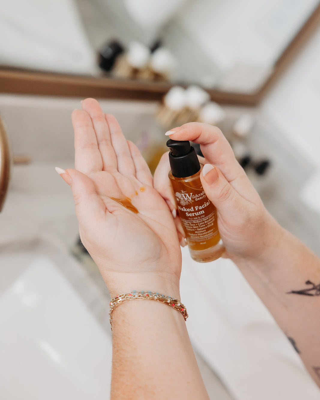 A person's hands holding a small amber bottle of hair oil with a black pump, with some of the oil visible on their palm, in a bright bathroom setting.