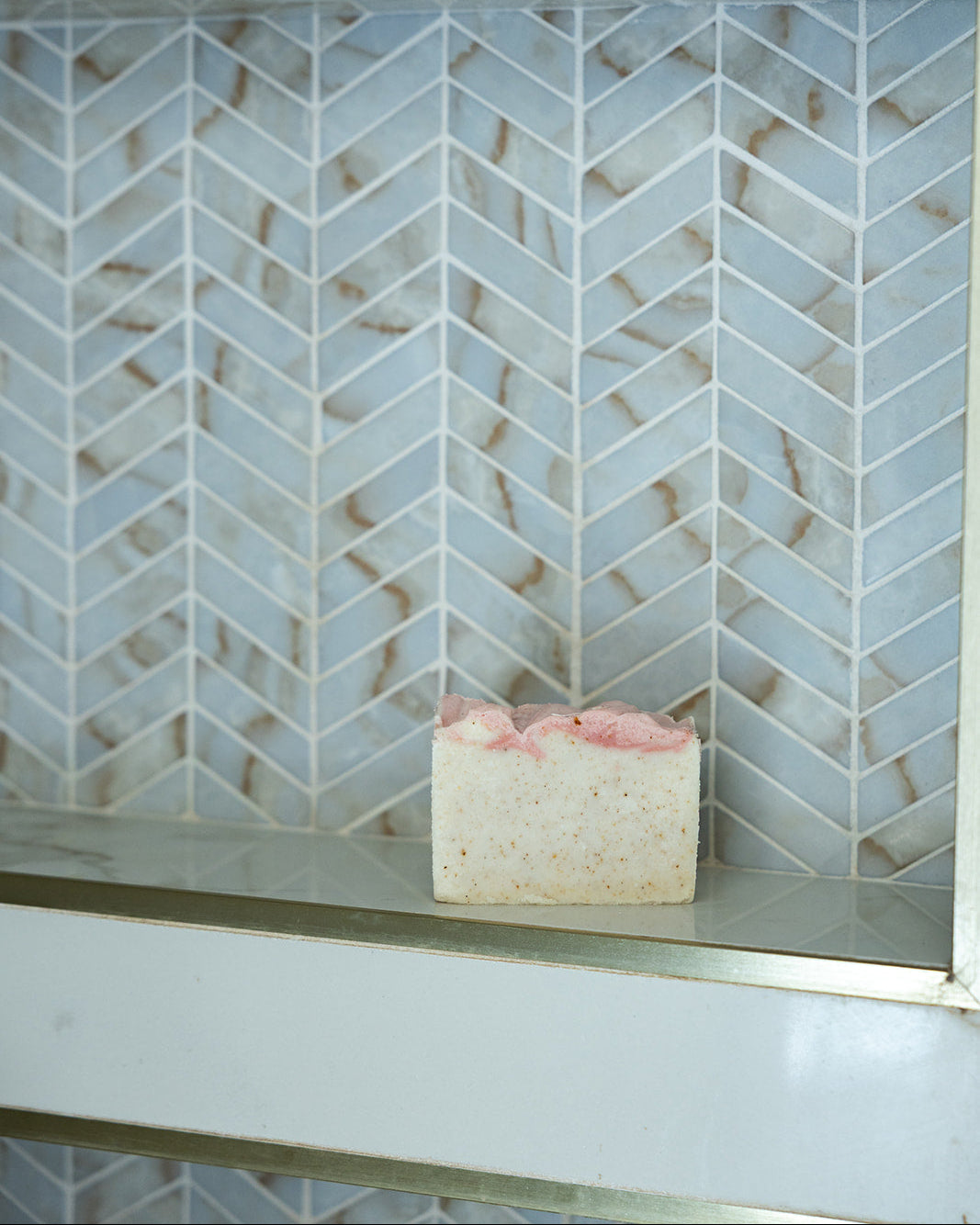 A rectangular bar of pale pink and white speckled soap rests on a glass shelf in front of a white and light gray herringbone tile backsplash with gold-toned metal trim.