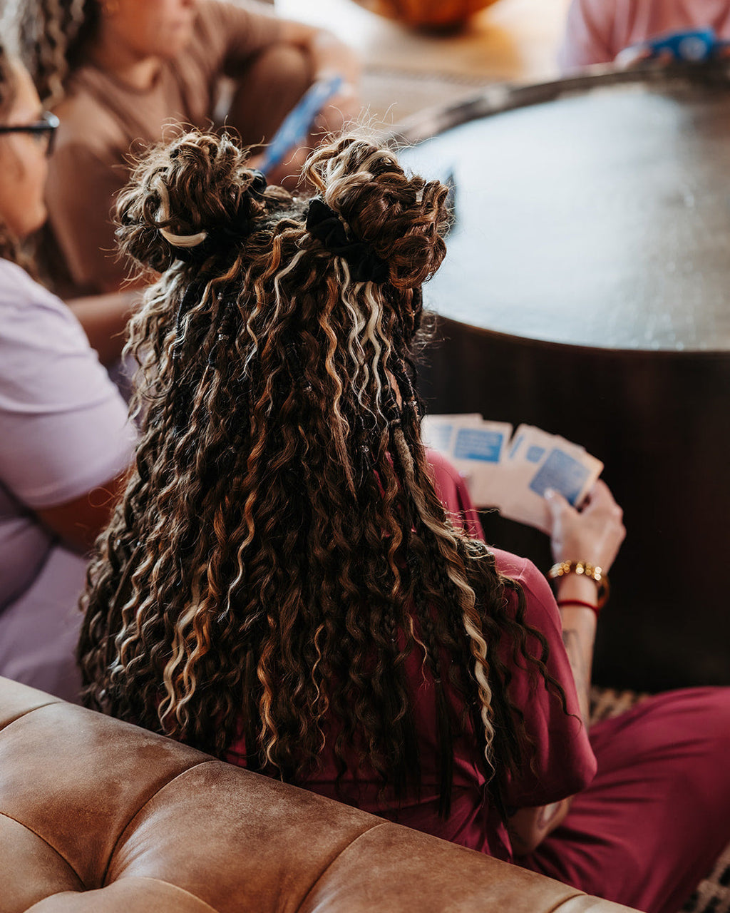Person with SHORTIES™ Braid-in Hair Extensions sitting on a couch, holding playing cards, with a pumpkin in the background