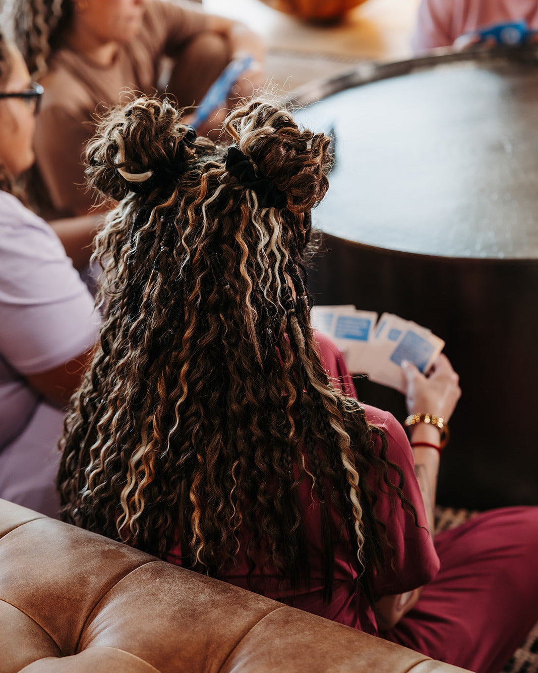 Person with SHORTIES™ Braid-in Hair Extensions sitting on a couch, holding playing cards, with a pumpkin in the background