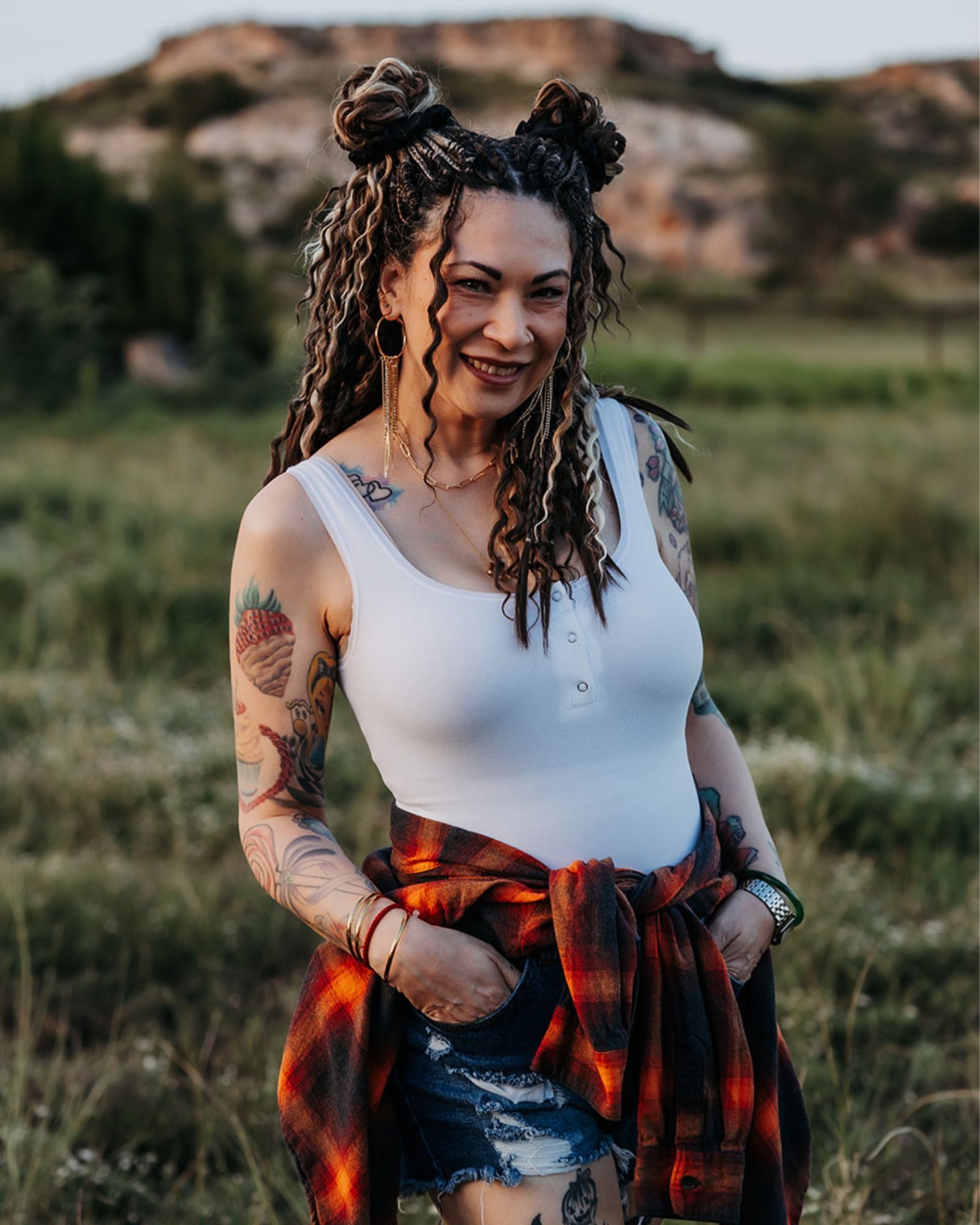 Woman wearing SHORTIES™ Braid-in Hair Extensions with tattoos standing in a field wearing a white tank top and plaid skirt.