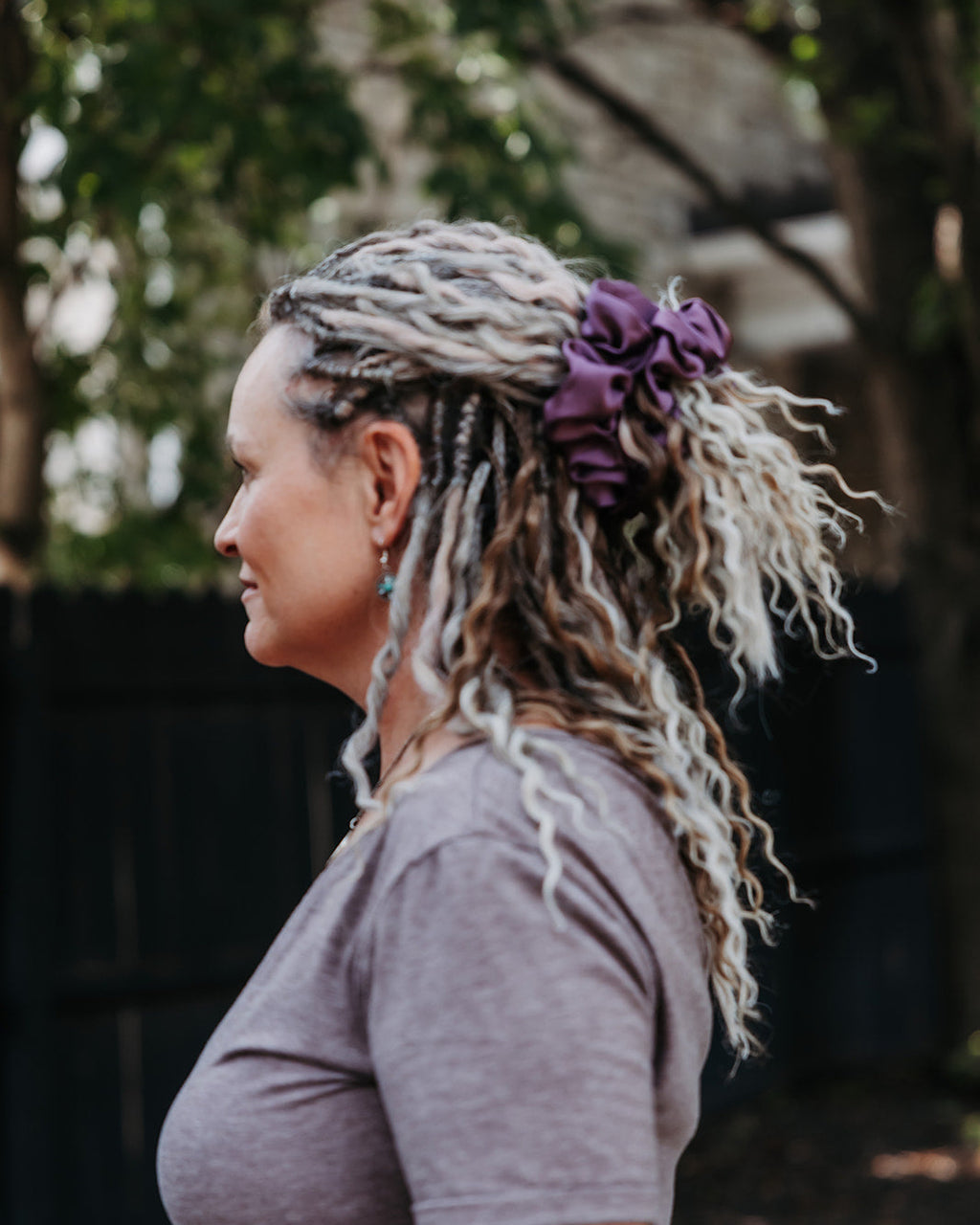 A woman in profile wearing a gray t-shirt and a purple satin scrunchie in an outdoor setting with trees in the background.