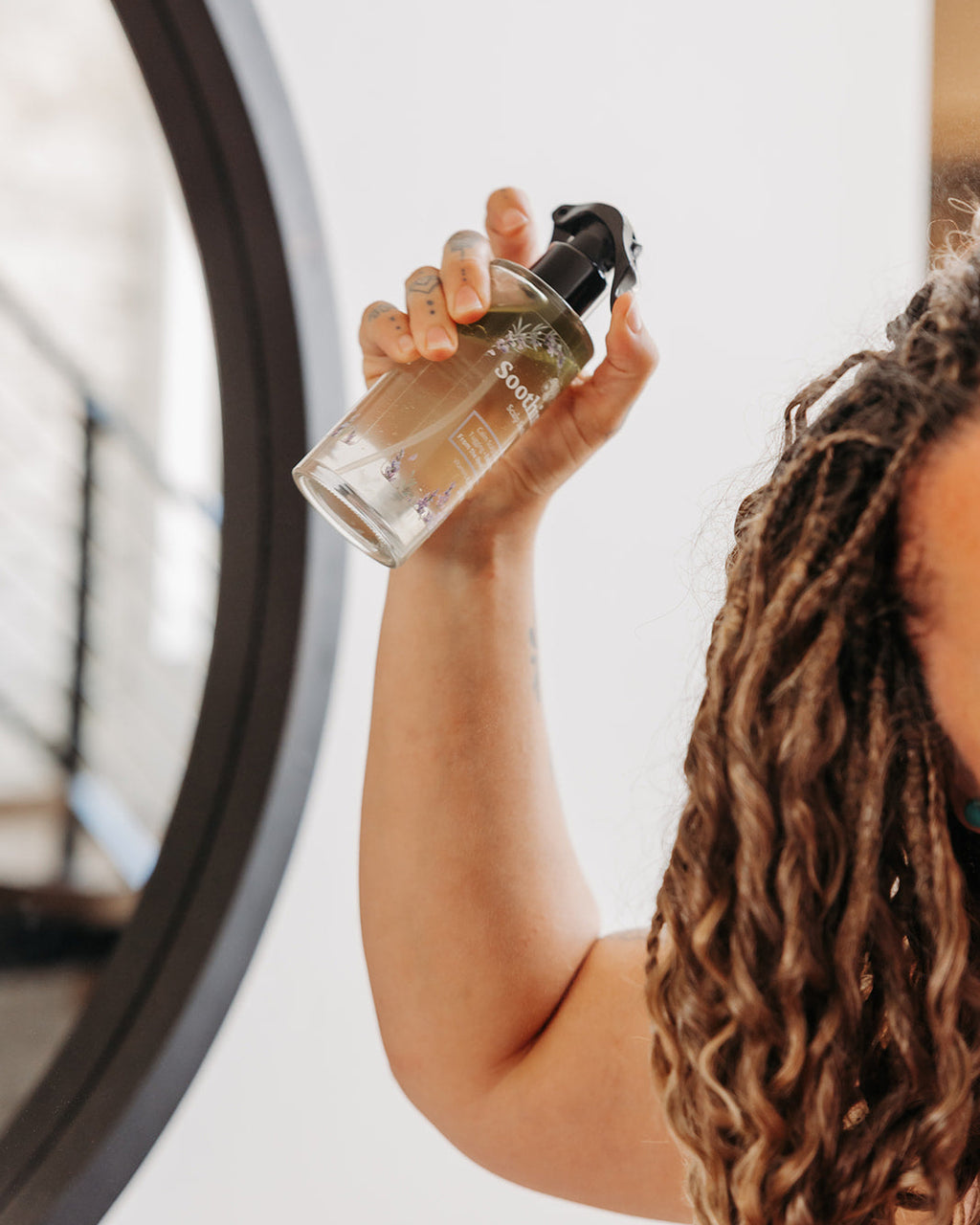 A person with wavy brown braid-in hair extensions holds a clear spray bottle with a black pump in front of their face, with a large round mirror visible on the left side of the frame.