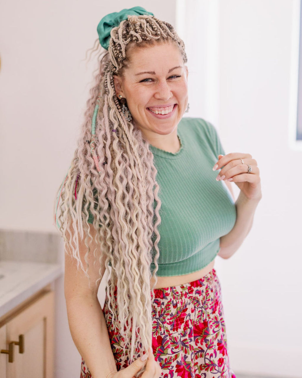 Woman with Venus Long Braid-in Hair Extensions wearing a green top and floral skirt in a kitchen.