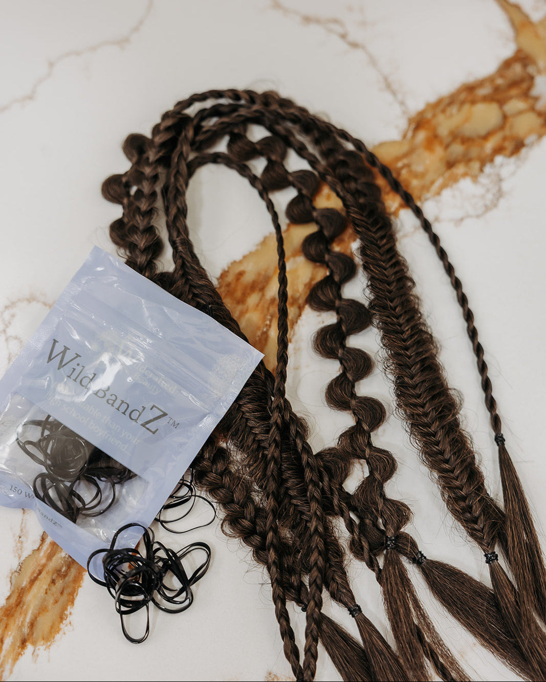 Dark brown braided hair extensions with various styles displayed next to a small clear plastic bag of black elastic hair ties on a white marble surface.