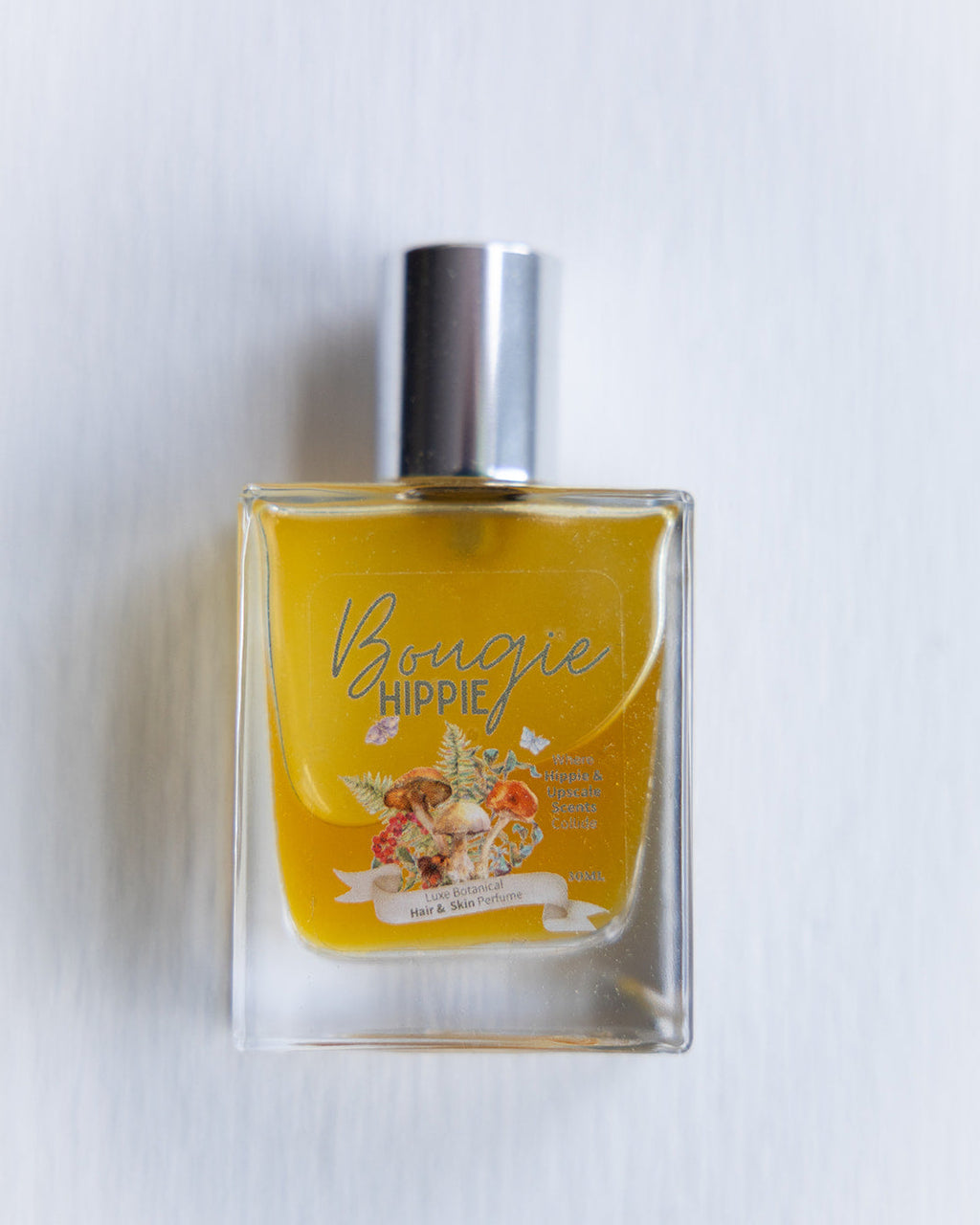 A clear glass bottle of Bougie Hippie Hair & Skin Perfume with a yellow-tan colored liquid visible inside. The perfume bottle is labeled and situated against a light background.