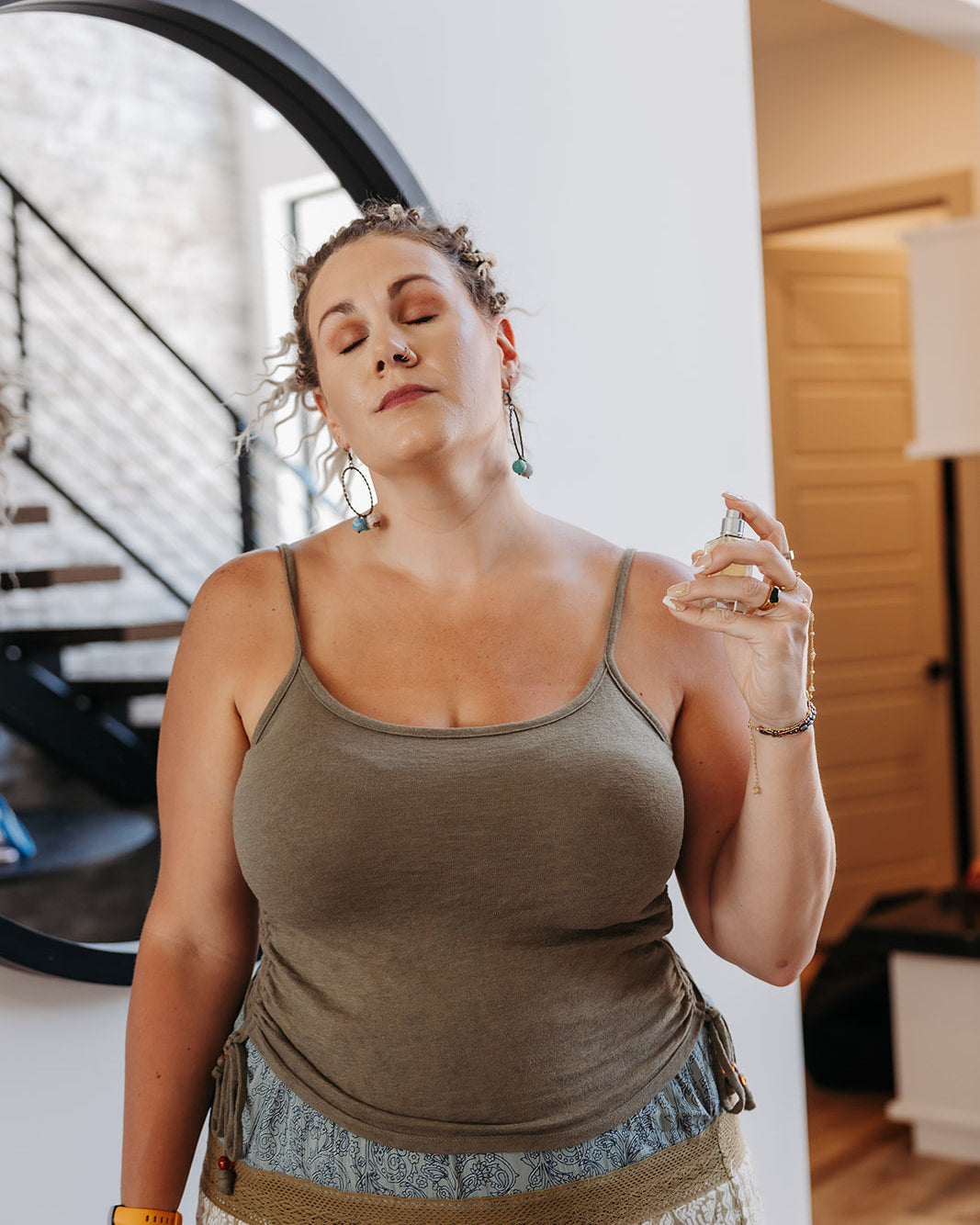 A woman with her eyes closed holds a small glass bottle near her shoulder, wearing a muted olive green tank top and a patterned skirt in an indoor setting with a staircase and mirror in the background.
