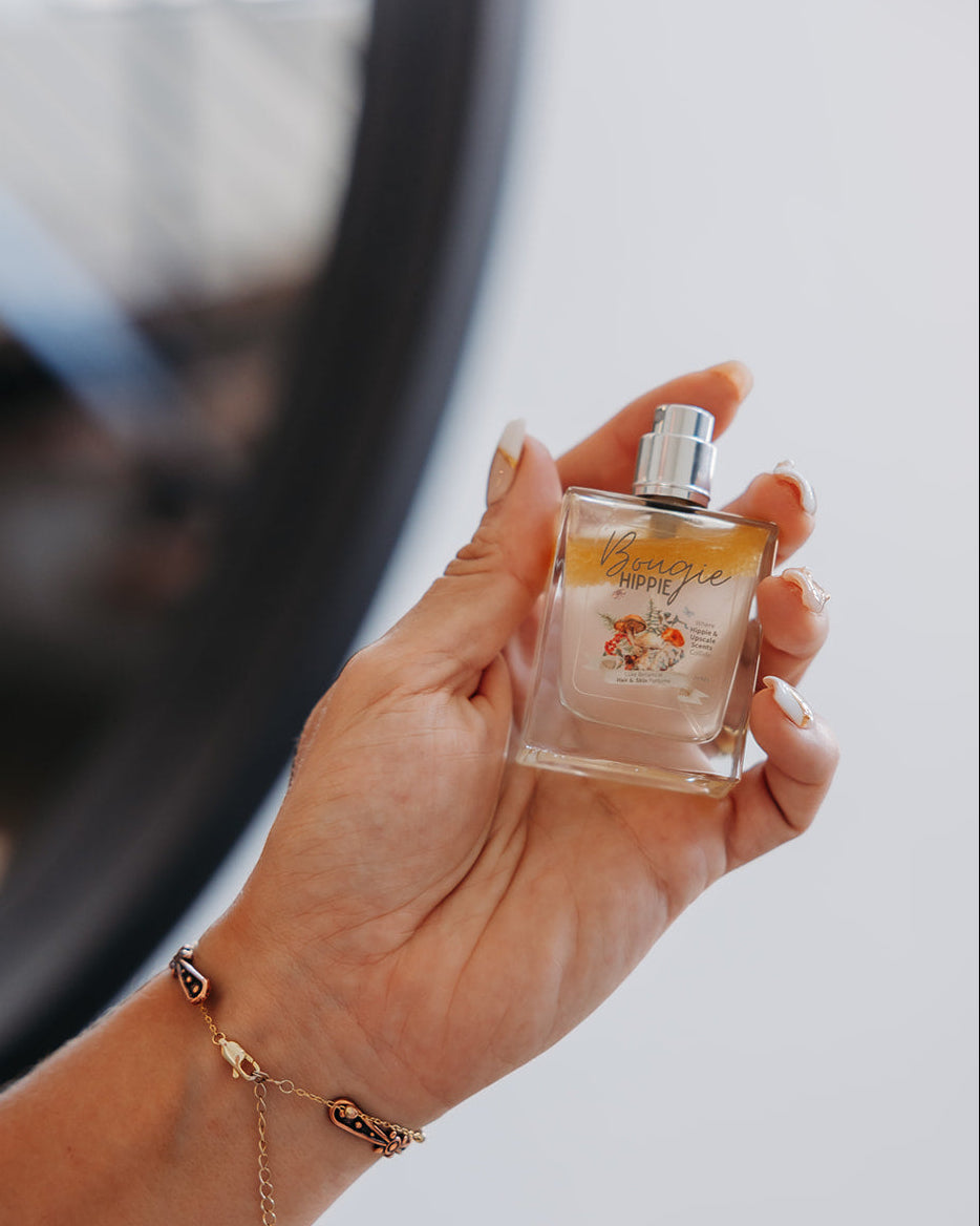 A hand holding a small glass perfume bottle with a silver spray top and a gold-toned floral label. The person is also wearing a delicate gold bracelet, and the background is softly blurred with a dark circular frame.