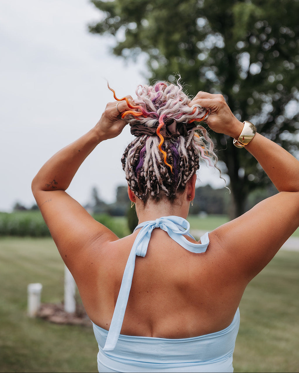 A woman viewed from behind outdoors, adjusting a messy bun hairstyle with dreadlocks in pink, purple, and range, with a brown scrunchie. She is wearing a light blue halter top.