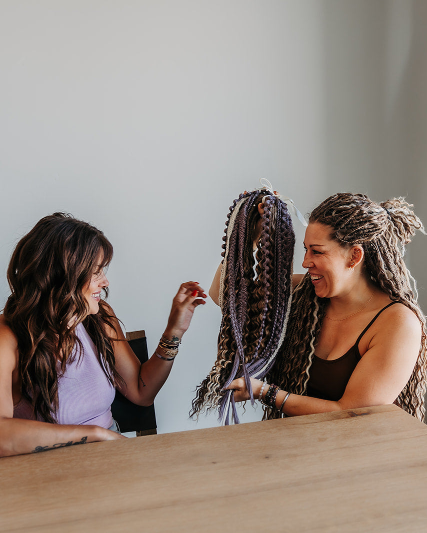 Two women smiling and laughing indoors near a wooden table, with one holding up a long, multi-colored braided hair extension to show the style. 