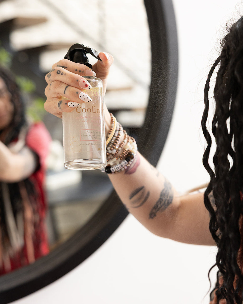 A person holds a clear spray bottle labeled 'Cooling Veil' in front of a mirror, with their hand, arm with tattoos and bracelets, and dark wavy hair visible. The reflection in the mirror shows their face.