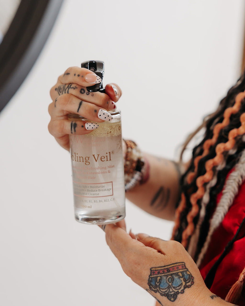 A person with tattooed hands and braided hair holds a clear glass bottle labeled 'Cooling Veil' against a plain light background.