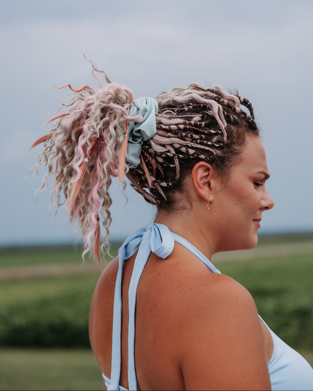 A woman in profile outdoors wearing a light blue halter top, with her hair styled in a high ponytail featuring pink and white streaks and a light blue scrunchie.