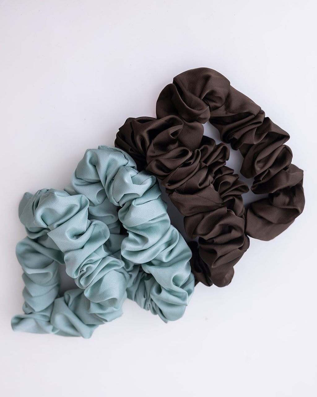 Four satin-like scrunchies, one in dusty teal and one in dark brown, arranged on a plain white background.