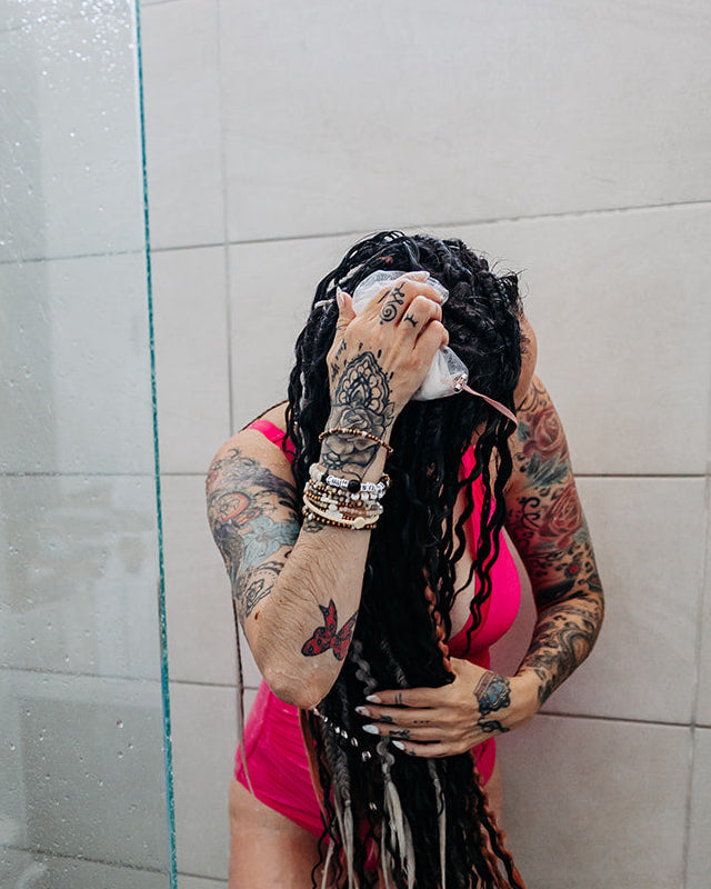 A woman with long black dreadlocks stands in a tiled shower, holding a Divinity Shampoo Bar to her hair. She is wearing a bright pink swimsuit, and the shower has a glass partition and dark tiled floor.