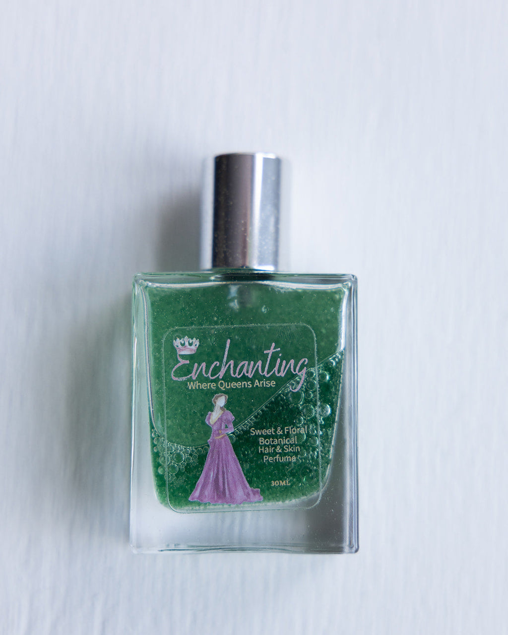 A clear bottle of 'Enchanting - Skin & Hair Perfume' with green liquid, displaying the product name and a silhouette of a woman in a dress.