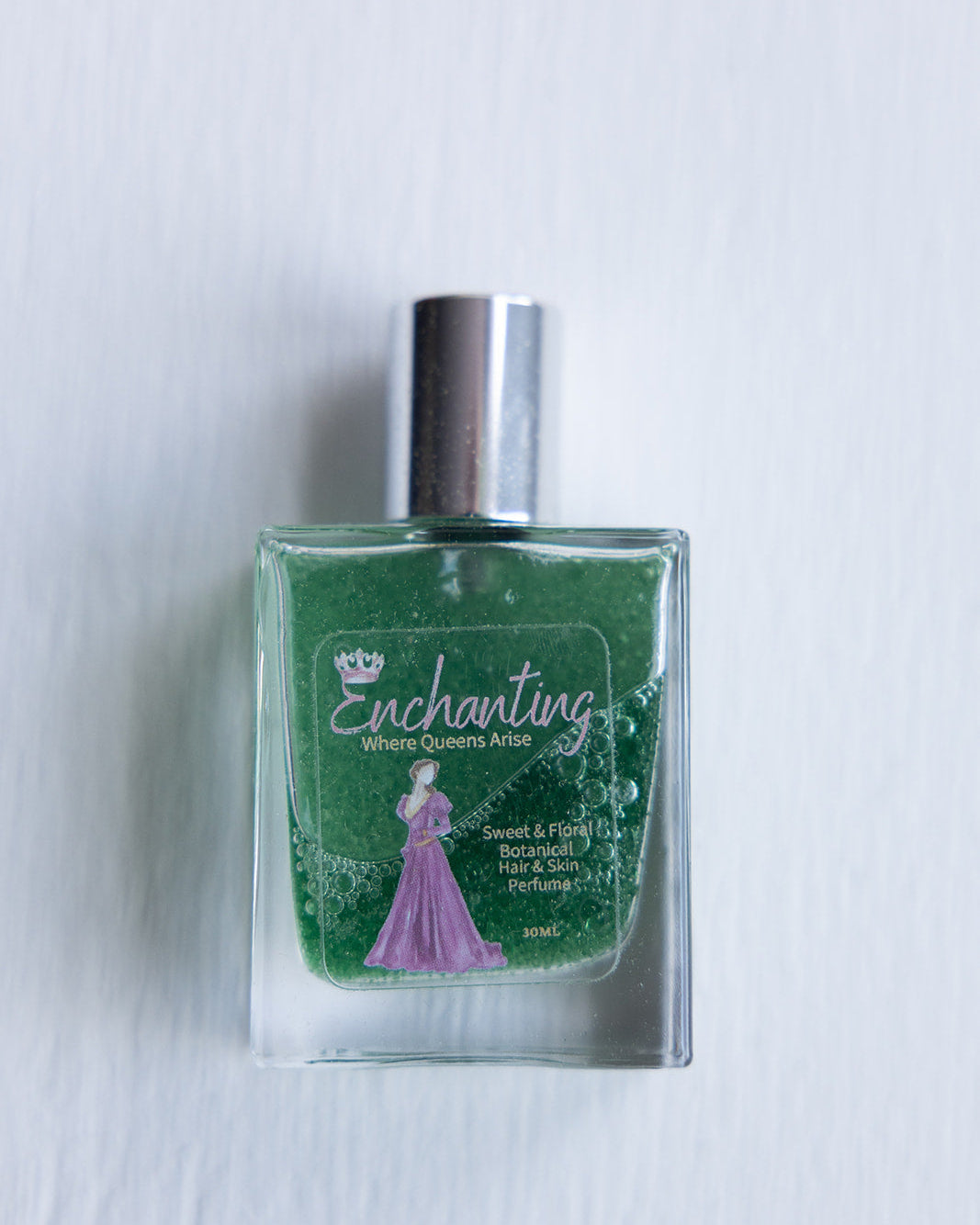A clear bottle of 'Enchanting - Skin & Hair Perfume' with green liquid, displaying the product name and a silhouette of a woman in a dress.