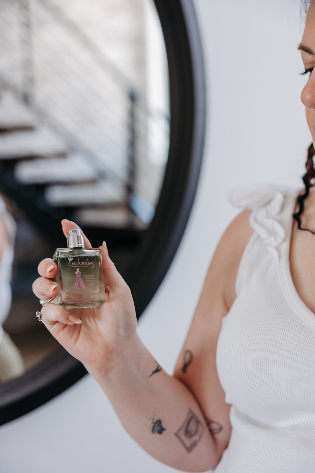 A woman in a white top holds up a small square glass perfume bottle with a silver cap. The bottle has green and clear liquid.