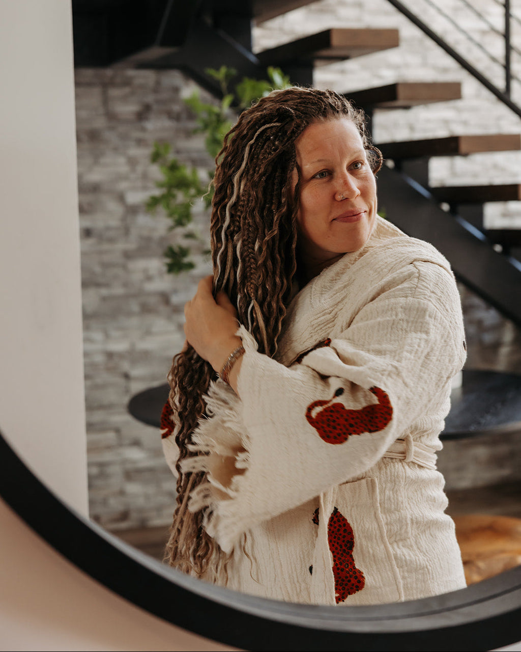 A woman with long, curly brown and blonde dreadlocks wearing a cream-colored knit cardigan with red accents, posing in front of a modern staircase.