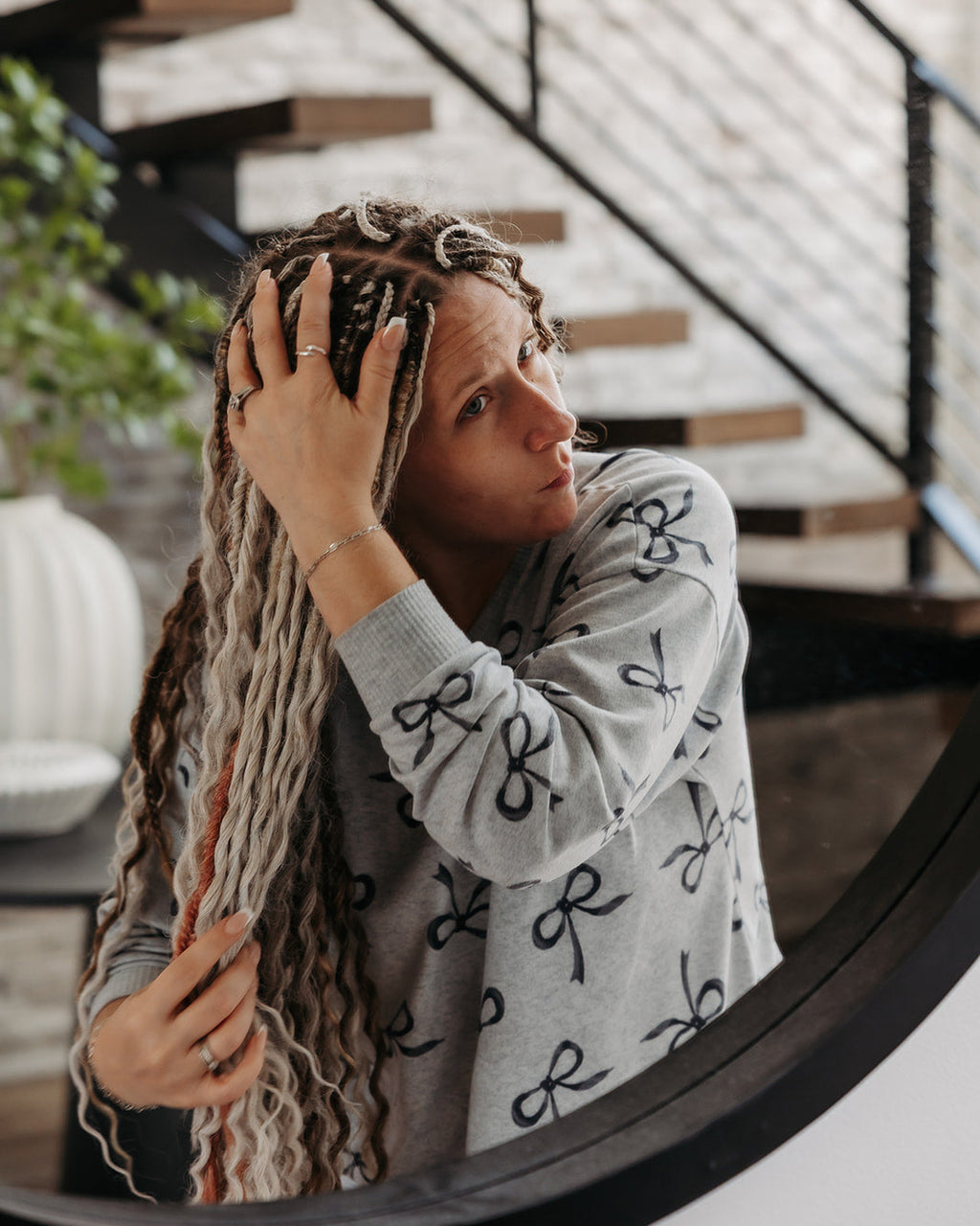 A woman with long, light brown and blonde wavy dreadlocks looks down at her hair while wearing a gray long-sleeve knit sweater with a black bow pattern. She is standing indoors in front of a staircase with wooden steps and a black metal railing.