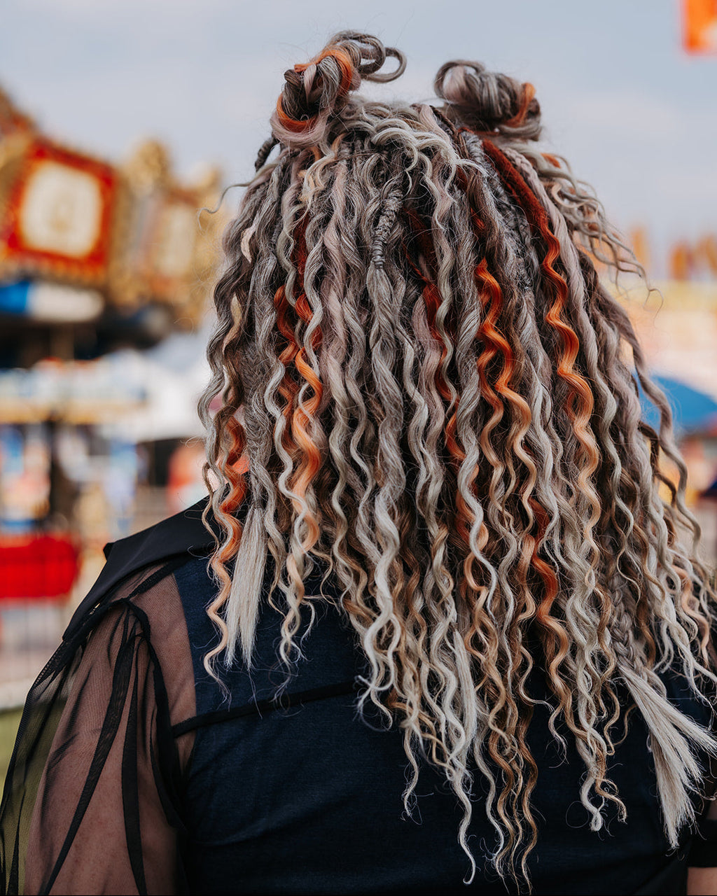 A person viewed from behind wearing a black top, featuring long, curly dreadlock-style hair in gray, white, and orange-brown tones. The background is a blurred outdoor fair or market scene with a blue sky.