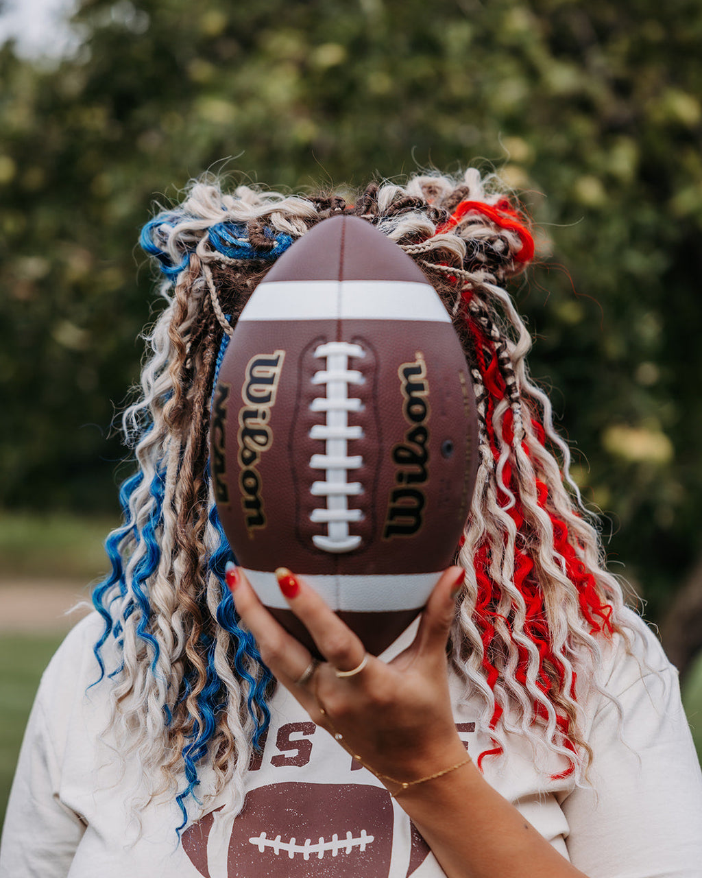 Girl with blonde, blue and red braid-in extensions outdoors holding a football in front of her face.
