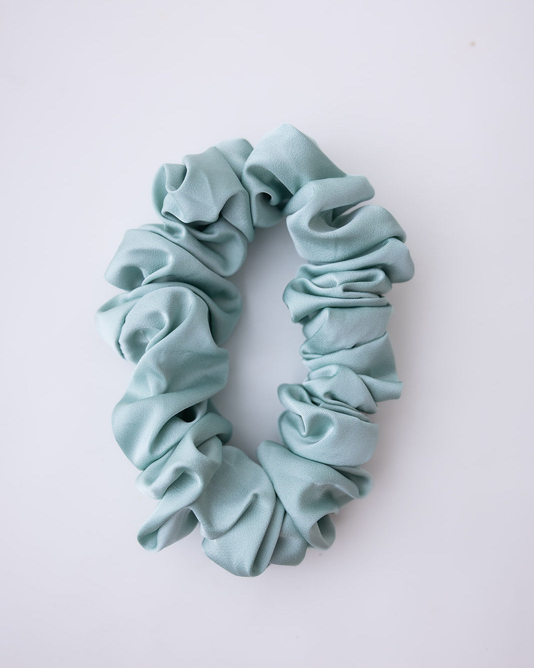 A dusty teal blue satin scrunchie with a gathered, ruffled texture, displayed on a clean white background.
