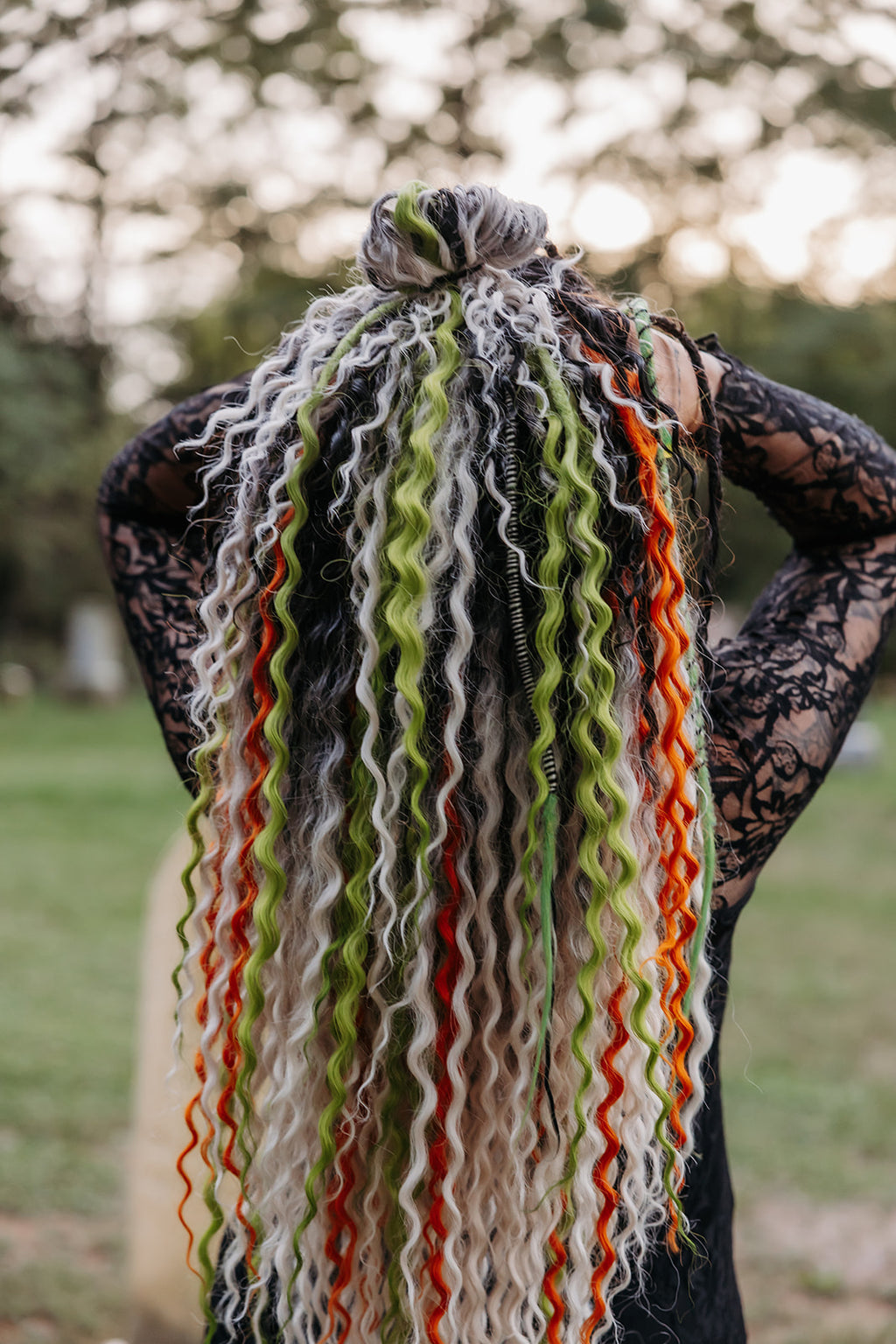 A person viewed from behind outdoors, wearing a black lace top and displaying a long, wavy dreadlocks with multiple colors including white to black, green, and orange.