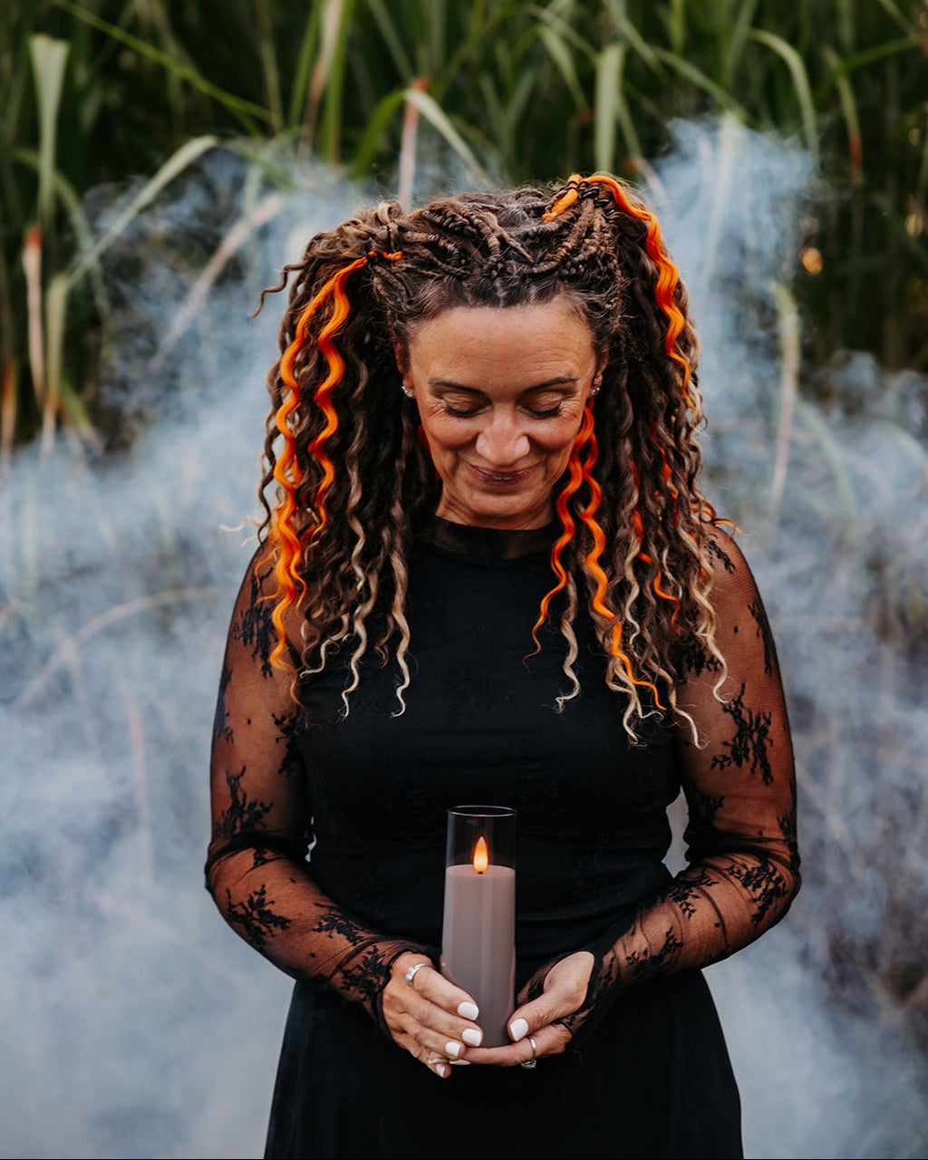 A woman with wavy brunette and orange dreadlocks stands outdoors in front of tall green reeds and smoke, holding a lit candle. She is wearing a black dress with sheer long sleeves featuring black lace patterns.