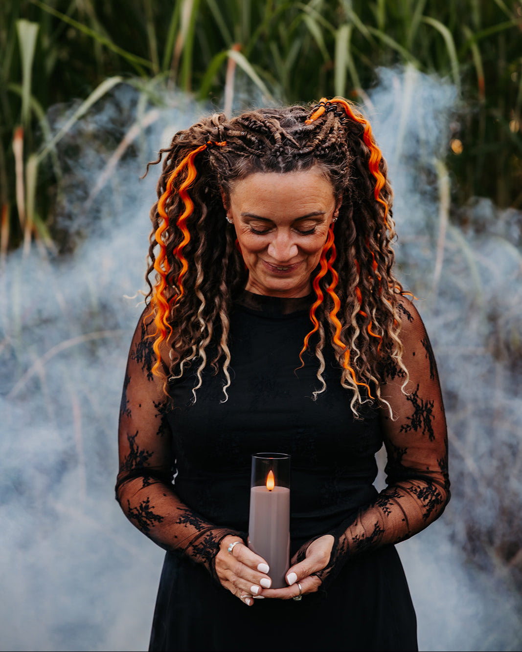 A woman with wavy brunette and orange dreadlocks stands outdoors in front of tall green reeds and smoke, holding a lit candle. She is wearing a black dress with sheer long sleeves featuring black lace patterns.