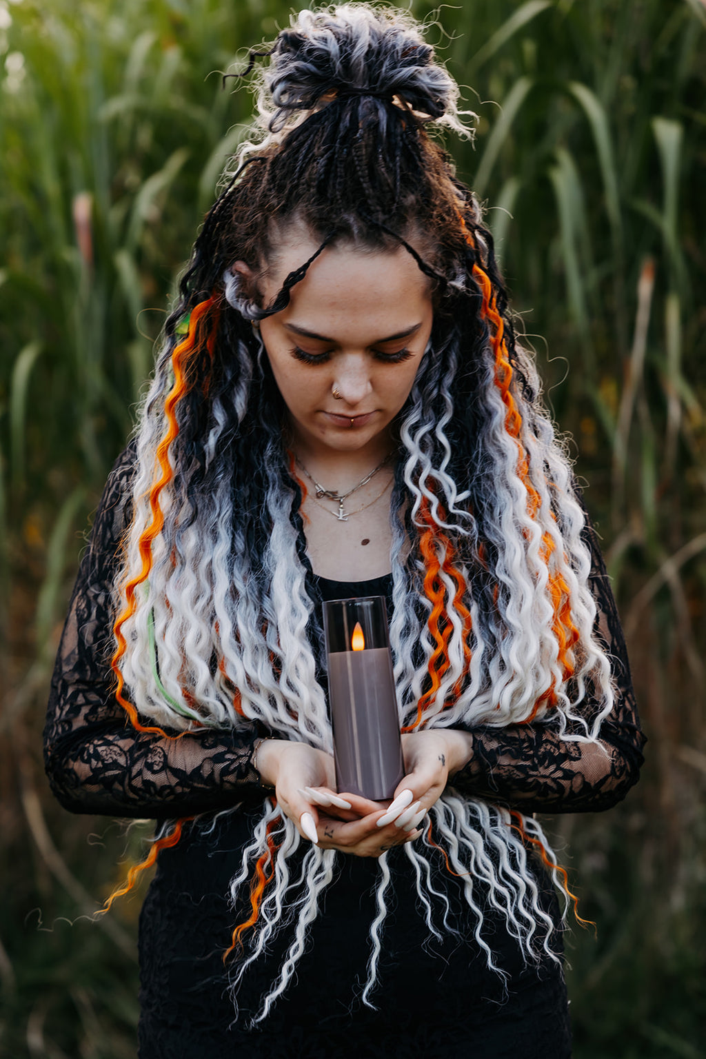A woman with long, curly black and white dreadlocks with orange accents holds a lit gray candle in her hands, wearing a black lace top, set against a blurred background of tall green reeds.