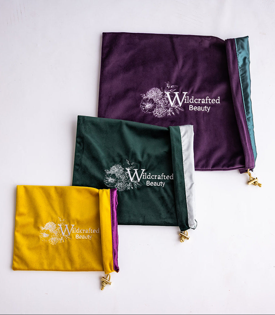 Three satin drawstring cosmetic pouches in purple, dark green, and mustard yellow, arranged diagonally on a white background. Each pouch features the 'Wildcrafted Beauty' logo printed in white, with gold-tone zipper pulls.