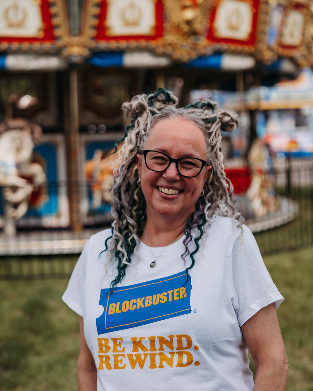 A smiling woman with glasses and gray dreadlocks stands outdoors in front of a blurred carousel, wearing a white t-shirt with a blue and yellow 'Blockbuster Be Kind. Rewind.
