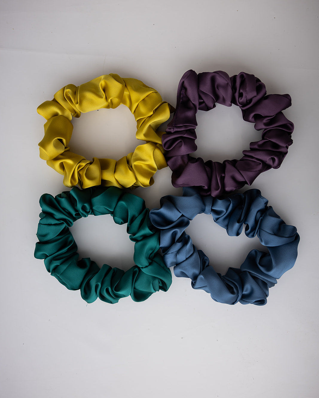 Four satin hair scrunchies in yellow, purple, teal, and slate blue, arranged in a 2x2 grid on a plain white background.